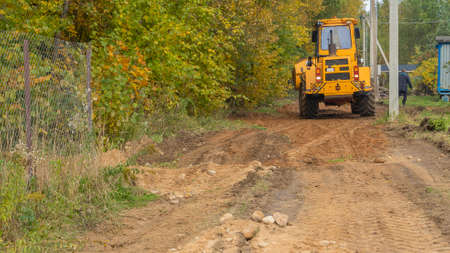 Rural road construction with grader in autumn daytime. The grader repairs the dirt road in the...