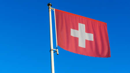 National flag of Switzerland on a flagpole in front of blue sky with sun rays and lens flare. Diplomacy concept. International relations. Space for text.の写真素材