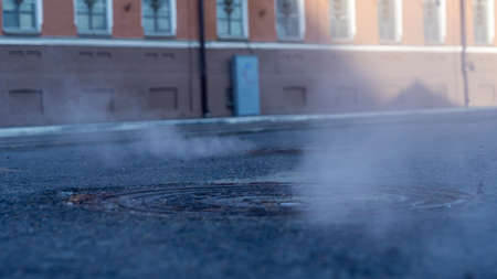 Manhole cover in a paved street on a cold day, as steam escapes from the sewer below up into the city street. Heavy steam coming out of a city center sewer.の写真素材