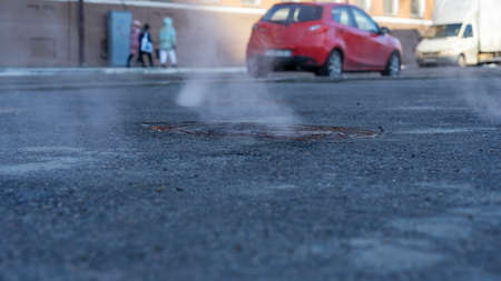 Manhole cover in a paved street on a cold day, as steam escapes from the sewer below up into the...