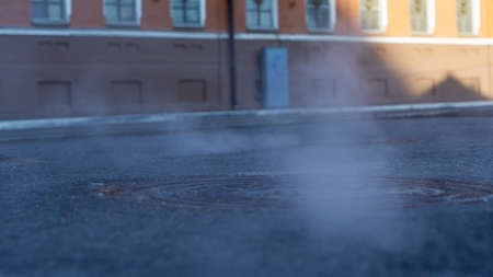 Manhole cover in a paved street on a cold day, as steam escapes from the sewer below up into the city street. Heavy steam coming out of a city center sewer.の写真素材