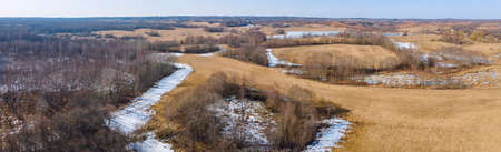 Aerial view of fields with copse, dry yellow grass, a stream, remnants of snow in early spring....