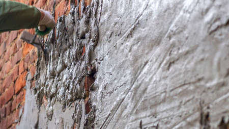 Plastering cement at wall for renovation house. Plasterer smoothing plaster on wall. Defocused background. Partly plastered old brick wall with damped plaster.の写真素材