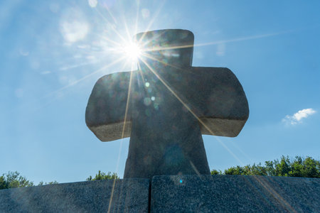 A solitary stone cross on a hill bathed in sunlight represents the enduring spirit of sacrifice and belief. Sun rays illuminate a stone cross. Stone cross under a clear blue sky.の写真素材