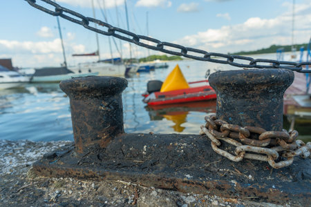 Vintage harbor scene with rusty bollard chain and boats under clear blue sky. Old maritime bollard with chain symbolizing strength and endurance at busy dock.の写真素材
