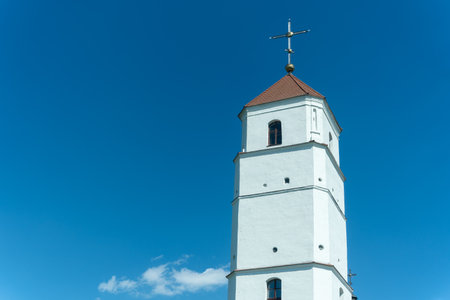 White bell tower with red roof and cross under clear blue sky. Historic church bell tower with red pyramidal roof golden sphere and cross rising against blue sky in religious architectural landscape.の写真素材