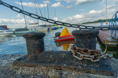 Vintage harbor scene with rusty bollard chain and boats under clear blue sky. Old maritime bollard with chain symbolizing strength and endurance at busy dock.の写真素材