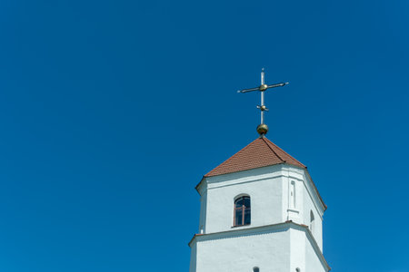 White bell tower with red roof and cross under clear blue sky. Historic church bell tower with red pyramidal roof golden sphere and cross rising against blue sky in religious architectural landscape.の写真素材