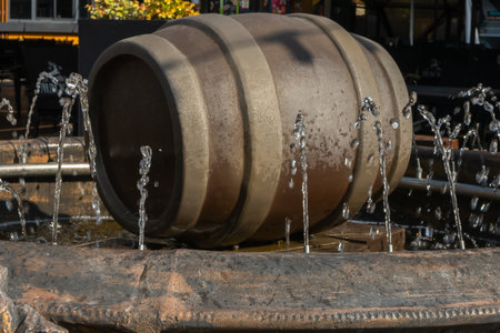 Water fountain with wooden barrel and circular jets in outdoor setting. Rustic barrel fountain with metal stand and water jets in public plaza with tables and chairs under natural daylight.の写真素材