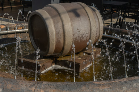 Water fountain with wooden barrel and circular jets in outdoor setting. Rustic barrel fountain with metal stand and water jets in public plaza with tables and chairs under natural daylight.の写真素材