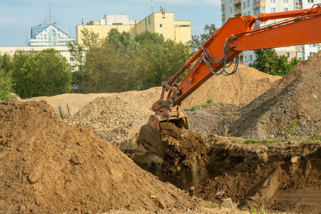 Excavator digging soil on construction site with urban buildings in the background. Industrial excavator performing earthmoving operations at construction site for residential infrastructure.の写真素材