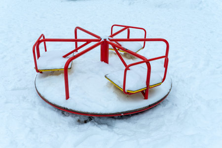 Red carousel covered with white snow on winter playground. Empty playground equipment in cold winter season with fresh snow on seats.の写真素材