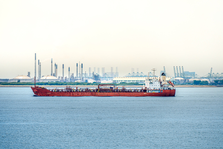 Red coloured hull large tanker sails along the coastal of industry port with large petroleum tank farm behide.の写真素材