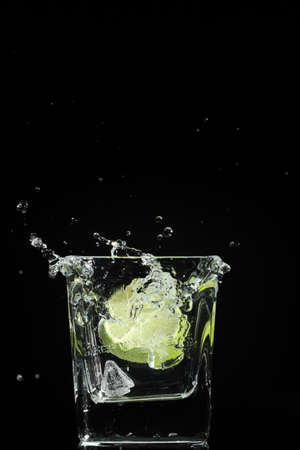 a splash of water in a glass with lime caused by an ice cube on a black backgroundの写真素材
