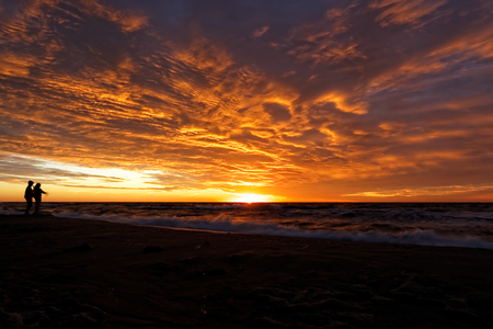 Sunset and dramatic clouds over the sea casts a glow on some indistinct human figures.の写真素材