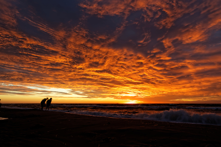 Sunset and dramatic clouds over the sea casts a glow on some indistinct human figures.の写真素材