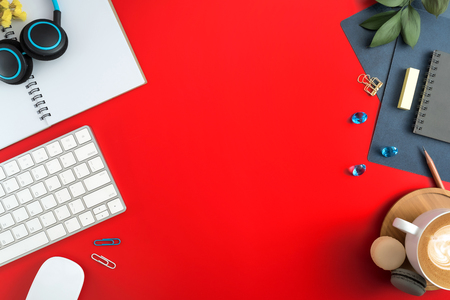 Flat lay, top view office table desk. Workspace with blank note book, keyboard, macaroon, office supplies, yellow flowers, green leaf, blue ornament and coffee cup on red background.の写真素材