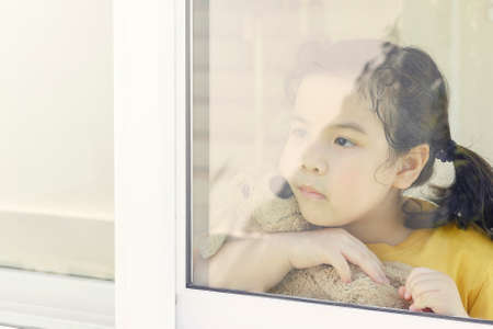 Little Asian girl stays home bored by school closings due to covid pandemic. Preschool girl looking outside her home window.の写真素材
