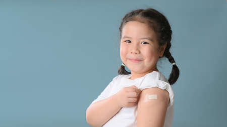 Coronavirus Vaccination Advertisement. Happy Vaccinated Little asian girl Showing Arm With Plaster Bandage After Covid-19 Vaccine Injection Posing Over Blue Backgroundの写真素材