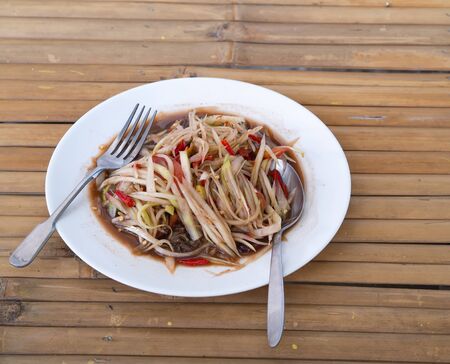 Hands hold folk and spoon. Eating Thaifood papaya salad with salted crab and fermented fish on fork and white dish, wooden table backgroundの写真素材