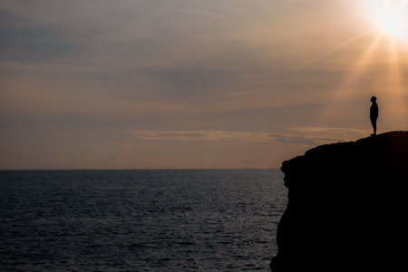 Silhouette of man stands on the edge of the abyss and looks at the sea on big stone at sunset in mountains.の写真素材