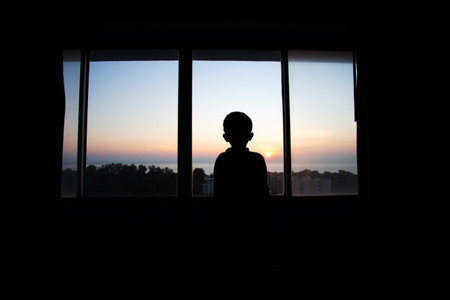 boy silhouette  standing and watching the morning sun rise in the sea on a chair in a windowの写真素材
