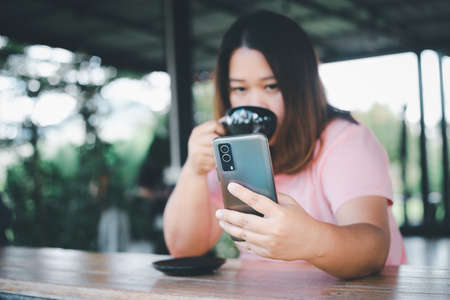 Asian woman holding and using mobile phone in coffee shop, enjoying the taste of coffee. or coffee shop with copy spaceの写真素材