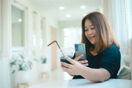 Asian female social media influencer taking photo of her black coffee before uploading to the internet. cheerful woman text messaging her friends in a modern and cute minimalist coffee cafe.の写真素材