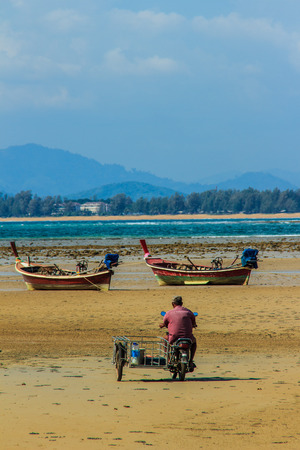 Fisherman was driving the motorcycle wagon to taken equipment to the boat in order to preparing his boat for the next fishing trip. His boat was park on the beach under blue sky and nearby the airportの写真素材