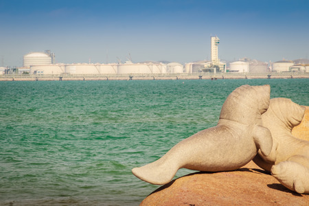 Lovely dugongs sculpture on Hadnamrin beach with blue sky and Maptaphut Industrial Estate in background at Ban Chang, Rayong, Thailandの写真素材