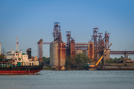 Old and rusty silos for rice storage before export. Floating crane is lifting the agricultural product to keep in silos and the ship is leaving the port to export.の写真素材