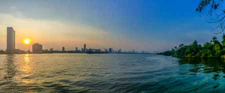 Panorama view of Chao Phraya river during sunset with high-rise condominium and buildings under construction in blue and yellow sky background. Silhouette riverfront view of real estate development.の写真素材