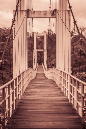 Vintage suspension bridge hanging across the river. Old lengthy hanging wooden footbridge with steel rails over river against cloudy sky background. Hanging wooden bridge at Kaengtana national park.の写真素材