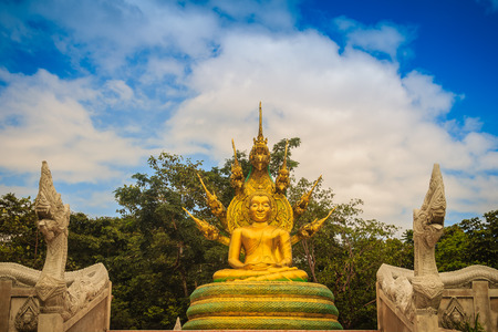 Beautiful golden Buddha statue with seven Phaya Naga heads under white clouds and blue sky background. Outdoor golden seated Buddha image protected by 7 heads Naga spreads cover on top in cloudy days.の写真素材