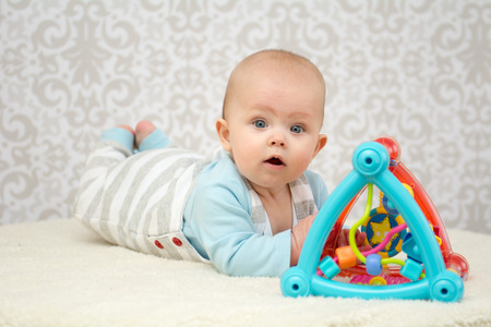 Blue eyes baby girl lying on belly on soft surface , playing with colourful toy and looking at the...