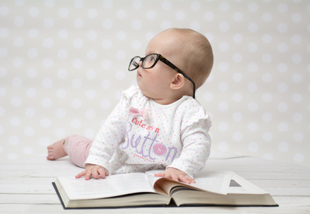 Funny portrait of a cute baby girl in glasses lying over a big bookの写真素材