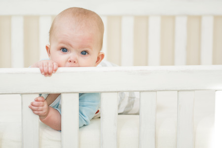 5 moths old baby girl bitting a white wooden crib.