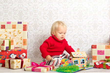 Dressed festively girl with stacks of present boxes around sitting on the floor and playing with paper gingerbread house.の写真素材