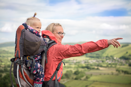Happy young woman trekking with  baby in a baby carrier and her a dog, pointing at something in the landscape. Hiking activity with child on family summer vacation, weekend nature tourの写真素材