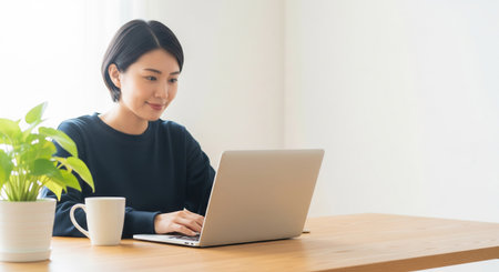 A twenties Japanese woman works on a laptop at a wooden desk in natural light. No UI or logos, a clean versatile home-work background.の素材