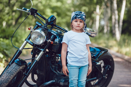Little girl in a white T-shirt and bandana standing near a motorcycleの写真素材