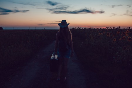 Girl in a cowboy hat standing with a suitcase on the road in the sunflower field. Waiting for help. Sunset.の写真素材