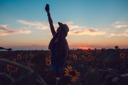 Girl in a cowboy hat lost in sunflower field catches a cellular phone. Sunset.の写真素材