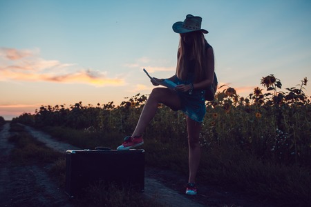 Girl in cowboy hat sitting on a suitcase in a sunflower field. Sunset.の写真素材