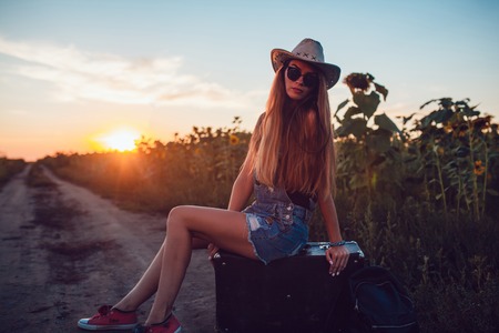 Girl in cowboy hat sitting on a suitcase in a sunflower field. Sunset.の写真素材