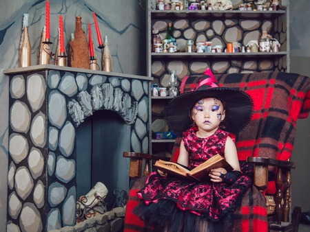 Girl in witch costume and make-up on her face sitting in a chair with a book in his hands in halloween decorationsの写真素材