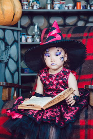 Girl in witch costume and make-up on her face sitting in a chair with a book in his hands in halloween decorationsの写真素材