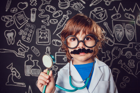 Happy little boy in doctor costume holding sthetoscope on dark background with pattern.の写真素材