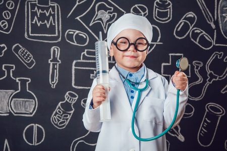 Happy little boy in doctor costum holding syringe and sthetoscope on dark background with pattern. The child has glassesの写真素材
