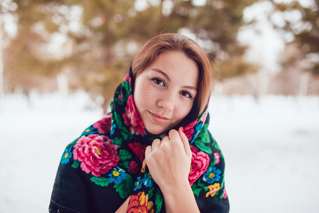Russian beauty woman with red hair and freckles in the national scarf stands on a background of trees.の写真素材